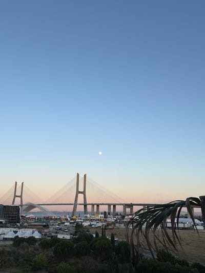 The image captures a wide-angle view of the Vasco da Gama Bridge spanning across the Tagus River in Loures, Portugal, during what appears to be the late afternoon or early evening, indicated by the soft light and clear sky with a visible moon. The bridge, a prominent cable-stayed structure, dominates the middle ground with its distinctive towers and suspension cables.
In the foreground, a sparse, dry landscape with scattered trees and bushes transitions into a more organized area that suggests a festival or event. This area is populated with white tents, portable structures, modular buildings, buses, and various vehicles. A large stage or structure with a black facade is visible on the left. A line of people can be seen walking or standing near the bridge's base, mingling around the event grounds.
The upper portion of the image is a clear, gradient blue sky, transitioning to a pale pinkish hue near the horizon, a common phenomenon during twilight. The moon is a small, white disc positioned in the upper center of the sky. A portion of a dried, brown palm frond extends into the bottom right corner of the frame, framing the scene from the right and adding a textural element. The overall scene depicts a confluence of infrastructure, nature, and human activity, likely an outdoor event set against the backdrop of a major landmark.