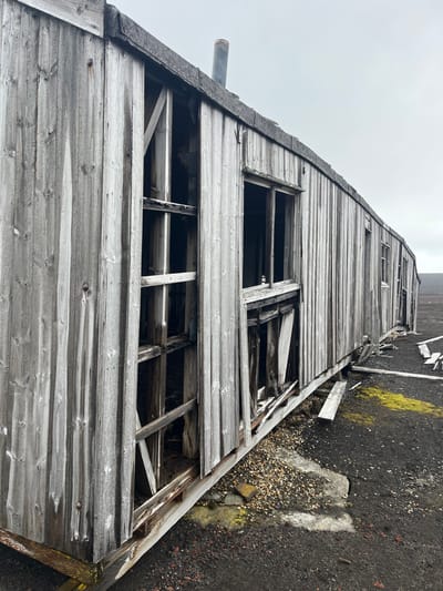 A dilapidated, gray wooden structure stands under a gloomy sky. Weathered planks form its exterior, revealing exposed framing and empty window spaces. The building appears abandoned, with signs of decay throughout. Patches of moss and dark soil surround its base, adding to the atmosphere of neglect and abandonment. A rusty pipe protrudes from the building's side. The structure seems to blend with the desolation of its environment.