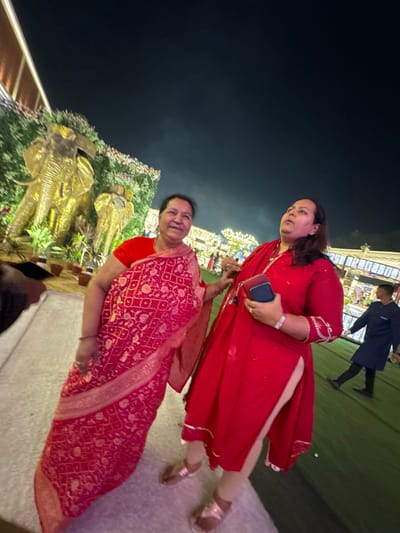 Two women in red attire stand outdoors at night. One wears a salwar kameez, holding a phone, while the other is draped in a sari. Golden elephant statues and floral arrangements illuminate the scene behind them. A dark-clothed person walks in the background. The lighting suggests a festive or celebratory event.