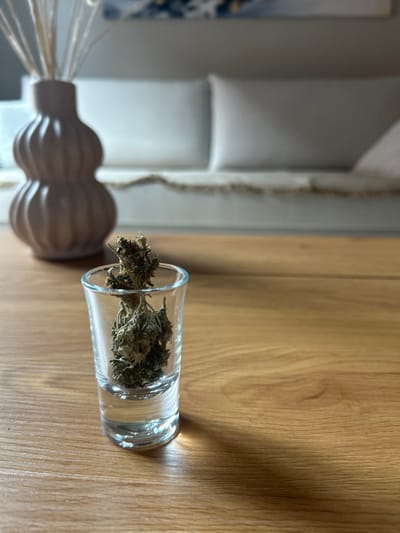 This image depicts an indoor scene. In the immediate foreground, a clear glass shot glass contains several dried, green-brown plant fragments, consistent with cannabis buds. The shot glass rests on a polished light brown wooden surface, likely a coffee table, which extends across the lower portion of the frame. In the left mid-background, out of focus, a light-colored, ribbed, two-tiered decorative vase holds several thin, dried, pale-colored stalks. Further in the background, a light-colored sofa with distinct back cushions is visible, also out of focus. A light-colored throw blanket with a decorative fringe is draped over the lower front of the sofa. Natural light illuminates the scene, casting a shadow from the shot glass onto the wooden table. The exact location within the city of Archanes Municipal Unit, Greece, is not identifiable from the visual information provided. No people are present, and no actions or interactions are depicted.