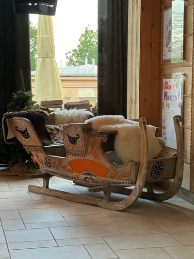 This is a medium shot, eye-level, indoor photograph of a decorative wooden sleigh. The sleigh is ornate, painted with orange and grey designs including floral patterns and a black star-like emblem. It is adorned with sheepskin rugs, one brown and one white, and appears to be set up as a prop for photographs.
In the background, a large, pale yellow umbrella is partially visible through a window. Outside the window, green trees and the rooftops of buildings can be seen. To the right of the sleigh, a wooden wall displays two framed posters. One poster, at the top, shows a green leaf and has text that is partially obscured but includes "Panczas". The lower poster is colorful and reads "Drogi Mikołaju!" (Dear Santa!) in large letters, with illustrations of stars and a cartoon figure.
To the left of the sleigh, a small, artificial Christmas tree is visible. The sleigh is positioned on a floor tiled with light-colored square and rectangular tiles arranged in a brick pattern. The lighting suggests an indoor space, possibly a cafe, shop, or guesthouse, given the decorative nature of the sleigh and the posters displayed. The overall atmosphere is festive and inviting. The presence of a Christmas-themed sleigh and decorations suggests a winter or holiday season setting.
The objects and context strongly suggest this scene is located in Zakopane, Poland, a popular tourist destination known for its wooden architecture and winter festivities, especially around Christmas. The poster "Drogi Mikołaju!" and the general aesthetic are consistent with Polish holiday traditions.