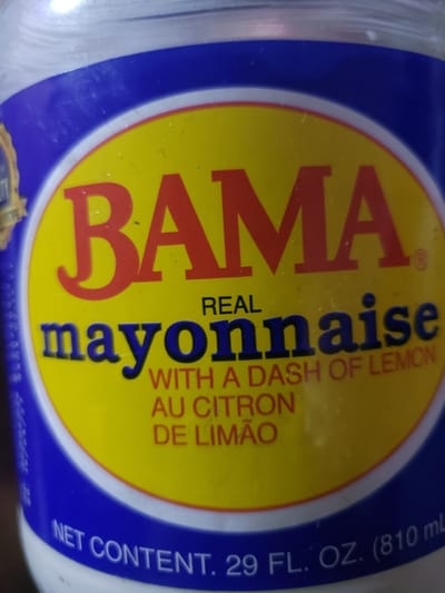 The image shows a close-up of a jar of BAMA Real Mayonnaise. The label is predominantly yellow and blue. Large red letters spell out "BAMA" at the top of the yellow section. Below this, in dark blue letters, is the word "mayonnaise". Smaller red text below that reads "WITH A DASH OF LEMON AU CITRON DE LIMÃO". Along the bottom of the label, in white text against a blue background, is "NET CONTENT. 29 FL. OZ. (810 ml)". The background is blurry and appears to be indoors, with some reflected light on the jar. There is no visible indication of Iganmu, Nigeria, or any specific city location.