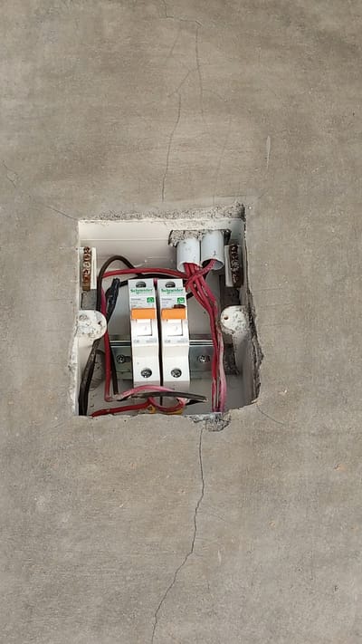 The image shows an electrical junction box embedded in a cracked concrete wall in Uteunreutoh, Indonesia. The junction box contains two Schneider Electric circuit breakers, labeled "C6". Red and black electrical wires are visible, connected to the circuit breakers and extending out of the box. There are also two white conduits protruding from the top of the box. The surrounding wall is rough and shows significant cracking. No people are present in the image.