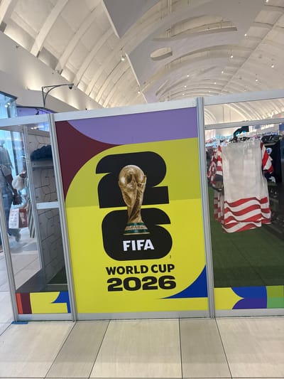 This image captures a promotional display for the FIFA World Cup 2026 situated within a large, brightly lit commercial interior, likely a shopping mall or airport terminal, in Sunrise, United States of America.

The central subject is a vertical display panel, framed in silver metal, showcasing the FIFA World Cup 2026 logo. The panel features a vibrant yellow background with a large black "26" graphic. Superimposed on the "6" is an image of the golden FIFA World Cup trophy. Below the trophy, the word "FIFA" is printed in white letters on a black rectangle, followed by "WORLD CUP 2026" in black text. The top section of the panel is purple, and a maroon band separates it from the yellow. The bottom border of the panel contains abstract colorful shapes in maroon, yellow, and blue.

To the immediate right of the display, a retail section is visible, featuring clothing items hung on racks. A prominent white sleeveless garment with red wavy stripes, resembling a patriotic or flag design, is displayed. The floor in this retail area is covered with green artificial turf. Further into the background, additional clothing and display units are visible.

To the immediate left of the display, a glass panel reflects part of the environment. A person wearing a light-colored shirt and blue jeans, carrying a red shopping bag, is reflected walking away from the viewer. Another partial reflection shows a person wearing a white hat or head covering. The floor in the foreground consists of light-colored square tiles with dark grout lines.

The expansive ceiling above is vaulted and white, constructed with a series of parallel, arched structural elements and visible cross-beams, characteristic of a large public space. Numerous small, circular recessed lights illuminate the area. A white dome-shaped security camera is mounted on a black, curved bracket extending from a wall segment in the upper left portion of the image. The overall lighting is bright and even.