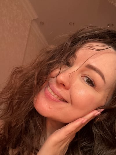 A close-up portrait shows a smiling woman with long, dark, and seemingly wet or damp wavy hair. She is angled towards the camera, looking directly at the viewer with a joyful and bright expression. Her left hand is gently placed on her cheek, near her ear, with manicured nails visible. Her skin appears fresh and radiant, and her eyes are bright. The background is a soft, uniformly light, warm-toned surface, possibly a wall or ceiling, featuring two discernible small, circular, recessed objects that could be lights or fixtures, hinting at an indoor setting such as a bathroom. The lighting is soft and warm, contributing to a pleasant and intimate atmosphere. The woman's demeanor conveys happiness and approachability. No text is visible in the image. The location context for this media file is Yuzhny, Russia.