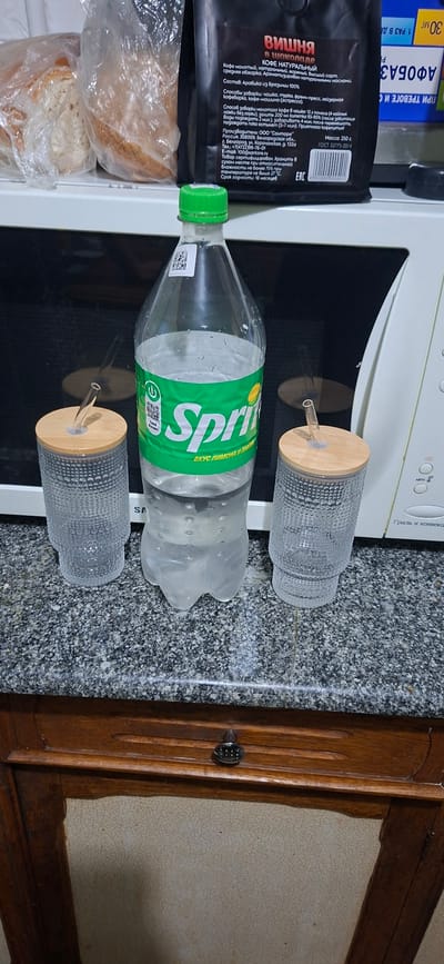 The image depicts a domestic setting, likely a kitchen, with a microwave oven dominating the background. In the foreground, a clear plastic bottle of Sprite lemon-lime soda is positioned centrally. To the left and right of the Sprite bottle are two clear glass jars with bamboo lids and glass straws, suggesting they are drinking glasses or containers. The countertop appears to be granite or a similar speckled material. Behind the microwave, a black package of coffee beans is visible, along with a clear plastic bag containing bread. The overall lighting suggests an indoor environment. There are no people visible in the image, and no actions are being performed. The scene is static and focuses on the arrangement of beverages and food items on a countertop. The specific location in Shakhtarsk, Ukraine cannot be determined from the visual information provided.
