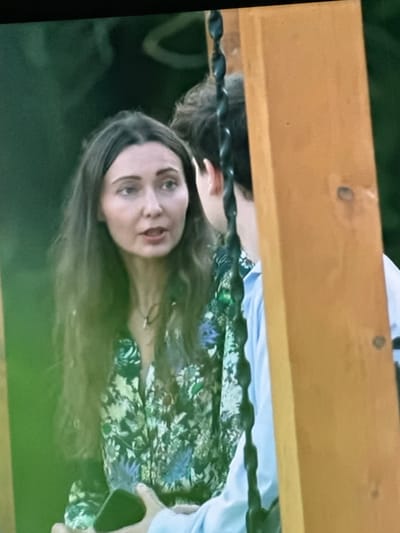 The image shows a close-up of two people, a woman and a man, in an outdoor setting. The woman is in the foreground, facing slightly to her right, with her mouth open as if speaking. She has long, dark brown hair and is wearing a floral-patterned dress. The man is to her right, with his back mostly to the camera, wearing a light blue shirt. A wooden beam, likely part of a swing set, is visible on the right side of the image, and a dark chain hangs to the left of the man. The background is blurred green, suggesting foliage. There is no visible indication of the city of Dondușeni, Moldova.