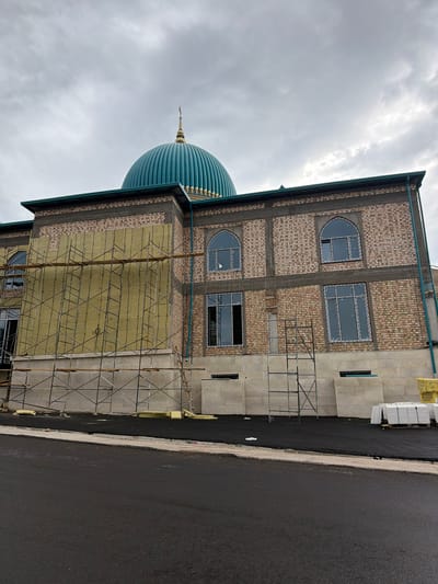The image shows the exterior of a mosque under construction in Khumsan, Uzbekistan. The building features a large, teal-colored dome with a golden finial, and is constructed with light-colored bricks. Scaffolding surrounds a section of the building where yellow insulation is being installed. Various windows, some arched and some rectangular, are visible on the brick facade. The ground in front of the mosque is covered in fresh asphalt, and a pile of white tiles on pallets is present to the right. The sky is overcast with gray clouds.