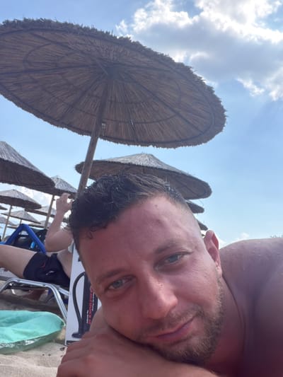 A low-angle selfie captures a man lying on a sandy beach under a bright sky in Logkari, Greece. The primary subject, a male with short dark hair, light eyes, and a light beard, is shirtless, resting his head on his right arm, and looking directly into the camera with a slight smile. His skin appears tanned. Above him, the underside of a large, circular straw beach umbrella fills the upper frame, with a blue sky and white clouds visible beyond its perimeter. To his left, in the mid-ground, several more straw umbrellas extend into the distance. A person wearing dark shorts is partially visible in the background, lying on a beach chair under one of these umbrellas, with their left hand raised near their head. On the sand to the primary subject's left, a white box for an "Intex Double Quick Air Pump" is partially visible, next to a deflated or partially inflated light green object, possibly a pillow or float, which rests near the metal frame of a beach chair. The overall scene depicts a sunny day at a beach location.