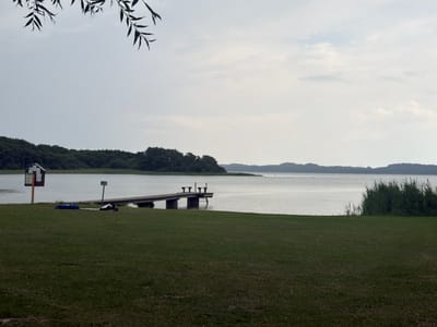 The image shows a wide shot of a lake and its surrounding landscape, with a wooden dock extending into the water. In the foreground, a grassy area slopes down towards the lake. On the left side of the grassy area, there is a wooden information kiosk. To the right of the kiosk, on the grass, are two blue items, possibly bags or mats, and a person lying down on a mat, facing away from the camera, dressed in dark clothing. Further to the right, a wooden dock with two benches at the end juts out into the lake. The lake is calm and reflects the overcast sky. On the far side of the lake, a tree-covered shoreline is visible, with hills in the distance. On the right side of the image, tall reeds grow along the edge of the water. The scene appears to be located in Köhn, Germany, at a lakeside recreational area.
