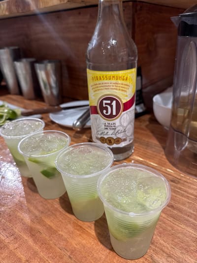 A bottle of "Cachaça 51" rests beside four plastic cups filled with a drink, likely caipirinhas, containing ice and lime wedges. The scene takes place on a wooden surface. The location is potentially Armação Dos Búzios, Brazil, given that cachaça and caipirinhas are common drinks in the region. The scene implies a social gathering or preparation for such, as multiple servings of the drink are ready.
