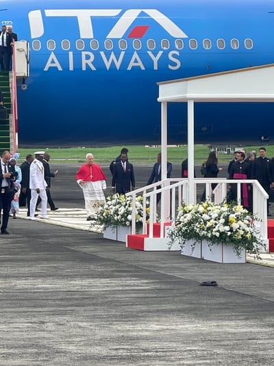 The image depicts an official ceremony taking place on an airport tarmac, likely in Malabo, Equatorial Guinea. A prominent blue passenger aircraft, clearly marked with "ITA AIRWAYS" on its side, forms the backdrop.

In the foreground, a portable white and red stairway, adorned with elaborate arrangements of white and yellow flowers, extends from the aircraft to the tarmac. A central figure, dressed in distinctive red and white religious vestments, stands near the base of this stairway, facing a gathering of people. This individual appears to be a high-ranking religious leader, such as the Pope.

Surrounding this central figure are various other individuals. Some, dressed in dark suits, including several wearing clerical collars, are visible on the stairway and further back on the tarmac. Closer to the viewer, a group of people in white attire, possibly ceremonial or traditional, are gathered on the tarmac, alongside others in suits. The activity suggests an official arrival or departure, with a formal welcoming or farewell party.

The scene is set during the daytime, under what appears to be an overcast sky, providing soft, diffused lighting. The overall mood is formal and respectful, befitting a significant diplomatic or religious visit.