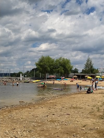 This image depicts a sunny day at a beach on the Slapy Reservoir near Radslav, Czechia. The scene is populated with numerous people engaged in various water and beach activities.
In the foreground, the sandy and rocky shoreline is visible, with some sparse vegetation. Further into the water, several individuals are present. Some are standing in the shallow water, while others are on paddleboards. A few people are actively participating in water sports, with some on paddleboards and others near small boats.
The midground of the image features a busy beach area. Numerous beach umbrellas, mostly yellow, are set up, providing shade for sunbathers. Several people are lying on towels or sitting on the sand. A small wooden building with a covered area and a few steps is situated on the beach, likely serving as a cafe or service point. Inflatable objects and various watercraft, including paddleboards and kayaks, are scattered on the sand and in the water.
The background of the image shows a dense line of trees along the edge of the reservoir, with a clear sky dotted with cumulus clouds. A harbor with many sailboats is visible on the left side of the image. A breakwater made of rocks extends into the water. The overall atmosphere is one of leisure and recreation on a warm day.