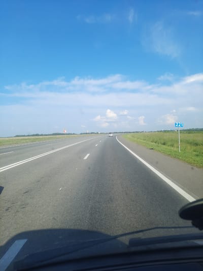 The image shows a highway scene from a first-person perspective, likely from inside a moving vehicle. The road stretches into the distance, marked with white lines. To the right of the road, there is a grassy verge. On the verge, a blue sign with the number "221" in white digits is visible. There are also some smaller warning signs with red and white markings along the roadside. The sky is a clear blue with scattered white clouds, suggesting fair weather. The sun appears to be casting a shadow from the vehicle on the right side of the image, and the general lighting indicates it is daytime. In the distance, the road is bordered by a line of trees, and some structures or vehicles can be seen far down the highway. The overall scene depicts a journey along a highway in a rural or semi-rural setting. The time of day is likely afternoon, given the shadows. The location context suggests this is in Pokrovka, Russia.