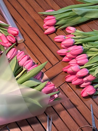The image presents an overhead view of a wooden surface, likely a table or floor, composed of rectangular planks arranged in a herringbone pattern, displaying varied brown tones. Scattered across this surface are numerous pink tulips and floristry tools.
On the left side, two partially assembled bouquets of pink tulips are visible. Each bouquet is wrapped in a combination of light pink paper and an outer layer of translucent, possibly light green or white, floral film. A roll of translucent wrapping film is positioned behind the upper left bouquet.
The right side of the frame is dominated by multiple bundles of unwrapped pink tulips, each featuring green stems and leaves, lying horizontally on the wooden surface. These tulips appear fresh and uniformly pink.
In the bottom left corner, a pair of silver-colored scissors with pointed blades is present. In the bottom right corner, another pair of similar silver-colored scissors is visible alongside a small spool of brown twine.
No individuals are depicted within the frame. The scene suggests an ongoing process of floral arrangement or bouquet preparation. The precise location within the city of Slavyansk-na-Kubani, Russia, is not identifiable from the visual information provided.