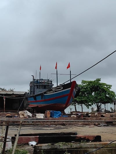 A large, blue and red fishing vessel, bearing the registration number 'TTH94899TS' and flying three Vietnamese flags, is dry-docked within a shipyard in Thuận An Ward, Vietnam. The vessel's hull is blue with a red bottom, a white stripe, and a black upper deck and superstructure; its bow is partially marked with the numeral '10'. It rests on wooden supports on an uneven concrete and dirt ground, which is littered with debris including crumpled white material, blue tarpaulins, wooden planks, and metal components. A dilapidated shed with a corrugated metal roof is partially visible to the left background. To the right, dense green trees stand, with a body of water faintly visible beyond them in the distance. A thick black cable extends diagonally from the upper right corner of the frame towards the vessel. The sky above is uniformly overcast and grey. No individuals are visible in the scene.