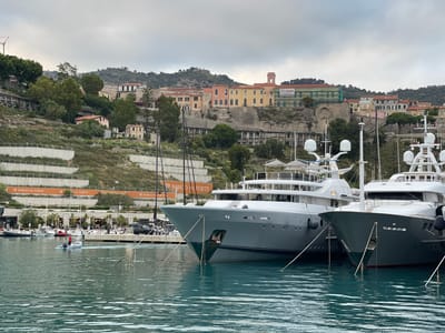 The image depicts a harbor scene in Ventimiglia, Italy, with several large yachts docked in the foreground. The water is a deep turquoise, reflecting the overcast sky and the modern vessels. Behind the yachts, a town is built on a hillside, characterized by pastel-colored buildings and terraced slopes dotted with greenery. Some of the buildings appear to be older, with stone walls and traditional architecture, while others are more modern.
In the middle ground, a person is operating a small inflatable dinghy, creating a wake in the water. A black SUV is parked near the waterfront promenade. The promenade itself has a row of buildings with outdoor seating, suggesting restaurants or cafes. Banners with the text "BORGO DEL FORTE" are visible along the waterfront.
The background shows a densely populated hillside with more buildings and vegetation, culminating in a ridge overlooking the town. The sky is cloudy, creating a diffused light that softens the scene. The overall impression is one of a bustling port town with a blend of old and new architecture nestled against a natural landscape.