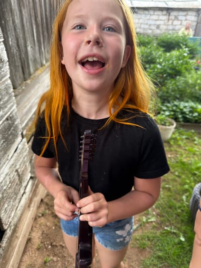 A young girl with bright orange hair and blue eyes is holding a dark-colored stringed instrument, possibly a ukulele or mandolin, which she is about to play. She is wearing a black t-shirt and denim shorts. The girl is standing outdoors, with a weathered wooden fence on her left and a lush green garden with a brick building in the background. The scene appears to be set in a rural or suburban area, potentially in the city of Bronnitsa, Russia. The overall impression is one of a candid moment, with the girl looking directly at the camera with a joyful expression.