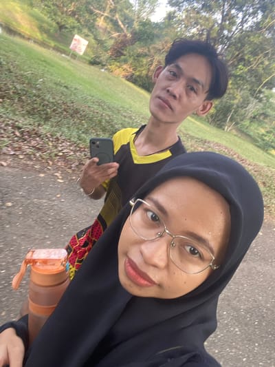 This media file is a selfie taken outdoors. The foreground features two people. On the right is a person wearing a black hijab and glasses, smiling at the camera. They are wearing a black top with a colorful patterned fabric visible underneath. An orange water bottle is also visible, held by this person. On the left, slightly behind and to the side, is a person with dark hair, wearing a black and yellow shirt. They are holding a smartphone in their hand.
The background shows a park-like setting with a paved path and grassy areas. Trees and foliage are visible, suggesting a natural environment. There is a sign on the left side of the path, though the text is not clearly legible. The lighting suggests it is either early morning or late afternoon. The people appear to be engaged in a casual outdoor activity, possibly a walk or run given the attire and presence of the water bottle. The overall mood seems relaxed and pleasant. The location context is Gudang Batu, Indonesia.