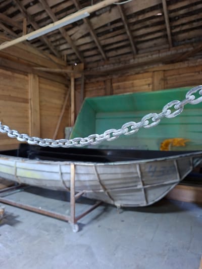 In a wooden shed in Žemaitkiemis, Lithuania, a metal chain hangs across the foreground, obscuring a partially visible boat. The boat, resting on a metal stand with wheels, is made of what appears to be aluminum and has the faded numeral "430" painted on its side. Behind the boat, a large, green plastic structure, possibly another boat or container, is visible. The shed's interior features exposed wooden beams and a sloped roof constructed of wooden planks. A fluorescent light fixture is mounted on a wooden beam overhead. The overall environment suggests a storage or workshop area for watercraft.