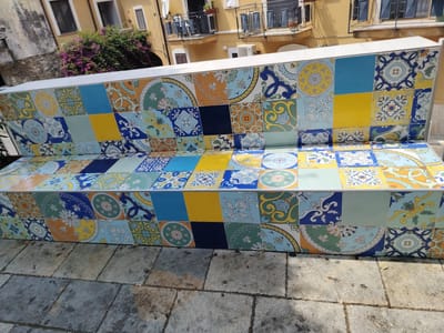 The image shows a decorative tiled bench in what appears to be an outdoor public space. The bench is covered in colorful ceramic tiles, many of which feature intricate geometric patterns and floral motifs in shades of blue, yellow, orange, green, and white. The tiles are arranged in a mosaic fashion, creating a vibrant and eye-catching design. The bench itself is a long, low structure, with a white top surface.

Behind the bench, a building with yellow walls and balconies with dark shutters is visible. Green foliage, possibly bougainvillea with bright pink flowers, spills over a wall on the left side of the frame. The ground in front of the bench is paved with large, irregular stone slabs, suggesting an older or traditional setting. The lighting suggests it is daytime, with bright sunlight casting shadows on the ground. The overall impression is one of a charming and picturesque location, possibly in a Mediterranean town like Agropoli, Italy, given the architectural style and vegetation. There is no visible text in the image. The scene is static, with no people or animals present, focusing entirely on the bench and its surroundings.