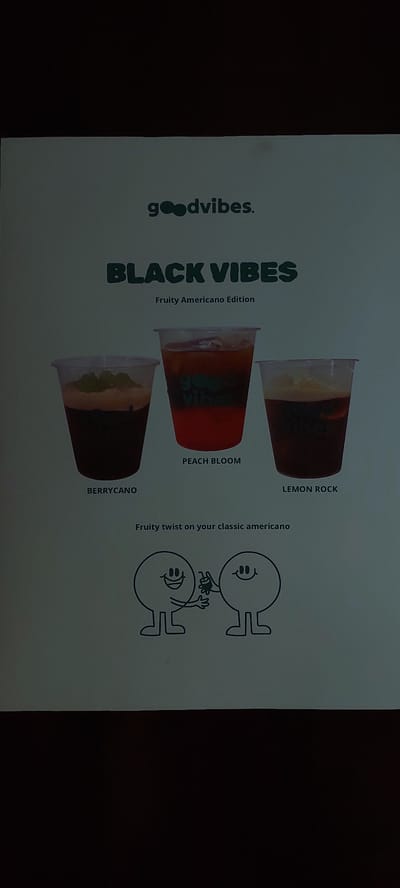 The image is a flyer or poster featuring three iced drinks in clear plastic cups, labeled "BERRYCANO," "PEACH BLOOM," and "LEMON ROCK." Above the drinks, the text reads "goodvibes. BLACK VIBES Fruity Americano Edition." Below the drinks, there is a tagline "Fruity twist on your classic americano." The bottom of the poster displays a simple line drawing of two round characters with arms and legs, one offering a small green object to the other. The overall presentation suggests a menu or advertisement for beverages, likely from a cafe or restaurant. The location of Pagedangan, Indonesia, is not visible or indicated within the image.