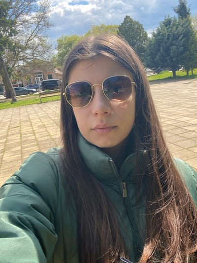 A selfie or close-up portrait captures a person outdoors in Kavarna, Bulgaria. The image, originally rotated 90 degrees clockwise, features the upper body of a young individual with long, straight dark brown hair that cascades over their shoulders. They are wearing a prominent green or teal-colored puffer jacket with a visible metal zipper pull. Gold-rimmed sunglasses cover their eyes, and the brand "Ray-Ban" is subtly visible on the left lens. The person's mouth is closed, showing a neutral expression.
The background is an outdoor setting under bright daylight, suggesting midday or early afternoon. Strong sunlight creates highlights on the hair, jacket, and the paved ground beneath. This ground appears to be a sidewalk or pathway made of light-colored, patterned paving stones. Beyond the paved area, there is green grass, various trees (some leafy, some with bare branches), and glimpses of distant residential buildings and parked cars. The sky is bright blue with scattered white clouds.