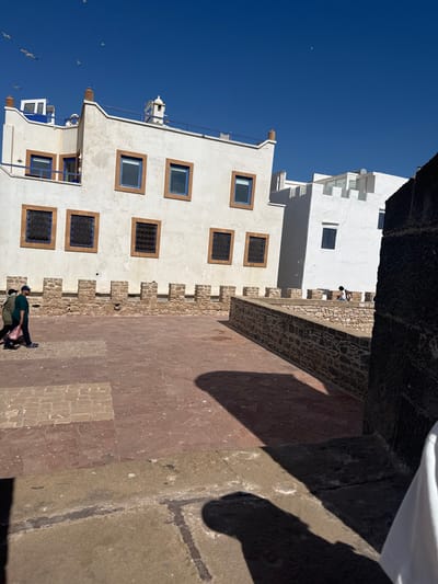 This is a medium-shot, eye-level, outdoor photograph taken in Essaouira, Morocco. The scene depicts a section of the historic city ramparts. In the background, two whitewashed buildings with blue-trimmed windows and brown decorative frames are visible. The building on the left has a white turret and a blue railing on a balcony, while the building on the right has a more modern, tiered design with numerous windows.
In the middle ground, two individuals are walking on a paved area. The person in the foreground, on the left, is wearing a dark green t-shirt, dark pants, and a dark baseball cap. They are carrying a dark bag in their left hand. The person behind them is wearing a light-colored jacket and dark pants. They are walking from left to right.
The foreground is dominated by a textured pavement made of reddish-brown and tan square and rectangular stones. A low stone wall, topped with crenellations, separates the walkway from a lower level. A shadow of a person or object is cast across the pavement from the right side of the frame. A dark, rough-textured stone wall is partially visible in the extreme right foreground.
The sky is a clear, bright blue with several seagulls flying in the upper left portion of the image. The architecture suggests the historical district of Essaouira, likely within the vicinity of the Skala de la Ville or similar coastal fortifications. The overall atmosphere is bright and sunny, indicative of daytime.