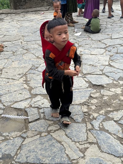 In Yên Minh Commune, Vietnam, a child wearing black pants, a black and orange long-sleeve shirt, and sandals stands on a stone pathway. A red carrier is wrapped around the child, holding a baby. In the background, there are glimpses of other people, including adults and children. The pathway is composed of large, irregularly shaped stones. A string of white objects lies near a small pool of water on the stone pathway.