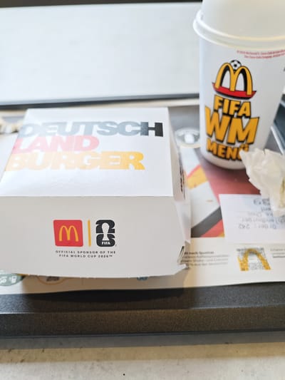 The image shows a McDonald's "Deutschland Burger" box and a promotional cup, suggesting a tie-in with a FIFA World Cup event. The box has "DEUTSCHLAND BURGER" printed on it in large, colorful letters, with "DEUTSCH" in gray and "LAND BURGER" in red and yellow, echoing the German flag colors. The cup features the McDonald's logo and text related to "FIFA WM" (World Cup) and "MENÜ" (Menu). Below the McDonald's and FIFA logos on the box, it states "OFFICIAL SPONSOR OF THE FIFA WORLD CUP 2026™". The setting appears to be a McDonald's restaurant, possibly in Tuttlingen, Germany, given the location context. The overall impression is a promotional display for a limited-edition menu item related to a sporting event. There are no people visible in the image. The lighting suggests it could be daytime, and the setting is indoors. Other visible text on a receipt includes "Order: 242".
