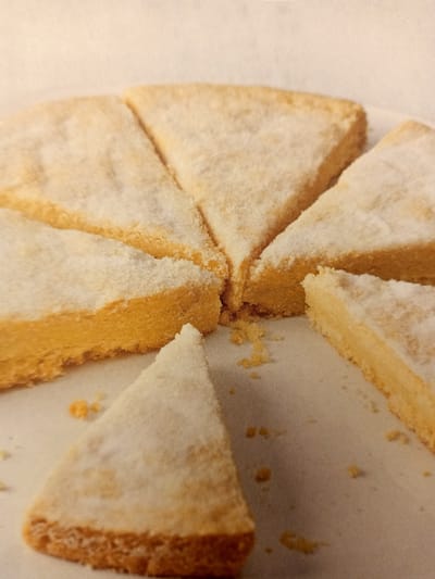 The image displays several wedges of shortbread cookies dusted with powdered sugar, arranged in a circular pattern on a textured surface. The cookies are golden brown with a slightly crumbly texture. Some small crumbs are scattered around the cookies. The background appears to be a light-colored, textured fabric. There are no people or any specific landmarks visible in the image that would allow for the determination of a location in Rumaithiya, Kuwait. The image is a close-up, studio-style shot of the food item.