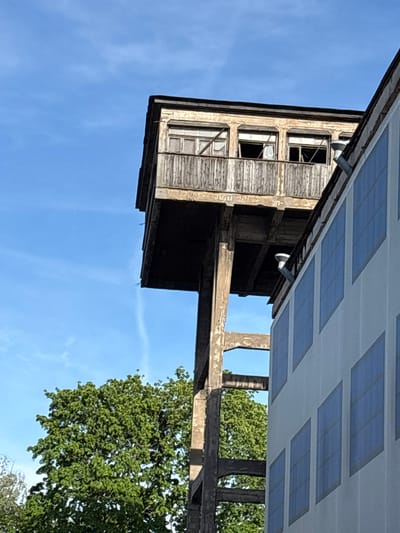 The image depicts an exterior view of a multi-story structure with weathered wooden siding and multiple windows, some of which appear broken. The building is supported by rough concrete columns. Above the structure, a modern roof-like element is visible, covered with a grid of solar panels. To the right, a lush green tree with dense foliage extends into the frame, with a clear blue sky providing the background. The scene is brightly lit, suggesting daytime, likely a sunny day. The overall impression is one of aged architecture juxtaposed with modern technology. There is no visible text in the image. The orientation of the image is such that the building appears to be hanging upside down, or viewed from an extremely low angle.