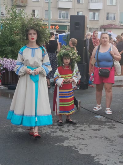 The image depicts a daytime outdoor gathering in Kryvdaivka, Russia, likely a festival or cultural event, given the traditional attire of two young girls. In the foreground, a young girl with a large, leafy floral wreath on her head and a colorful, patterned dress stands facing the viewer. To her left, an older girl, also adorned with a voluminous green floral wreath, wears a cream-colored, long-sleeved dress with blue embroidery and trim, and red shoes. Her expression is serious.
Behind them, a woman in a blue tank top and pink shorts, wearing white sandals, stands looking off to the side. A black crossbody bag is slung over her shoulder. Other people are visible in the background, including a woman in a red headscarf and a person in a black top and patterned skirt. A large black speaker is positioned to the left of the woman in the pink shorts. The setting appears to be a paved area in front of a multi-story building, with some greenery and flowers present. The lighting suggests it is daytime, and the weather appears overcast, though not actively raining. The mood seems to be one of community celebration. There is visible text on a building in the background, part of which appears to say "ET".