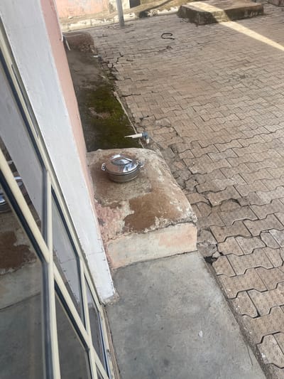 This media file depicts an outdoor scene, likely in Kporva, Nigeria. The primary focus is a paved ground area composed of interlocking concrete blocks, showing signs of wear and tear with some cracks and missing sections. Adjacent to this paving is a concrete structure, possibly a raised planter or wall, with a weathered and stained surface. Attached to this structure is a metallic water dispenser or tap, with a circular metal lid and a spout. A white pipe is also visible, connected to the water dispenser.

The foreground of the image is dominated by a section of a window or skylight with a beige frame and dark-tinted glass panes. The window structure appears to be at an angle, suggesting it might be part of a roof or a slanted wall. In the background, more of the interlocking block paving extends, and further along, there's a wall covered in green moss, indicating damp conditions or an area that doesn't receive much sunlight.

The lighting suggests it is daytime, with shadows cast across the paving. There are no people visible in the image. The overall impression is one of an aged, functional outdoor space, possibly in a residential or public area where water access is provided. The scene is static, with no activity or event clearly taking place. The condition of the paving and the concrete structure suggests a location that has experienced weathering and possibly has not undergone recent maintenance.