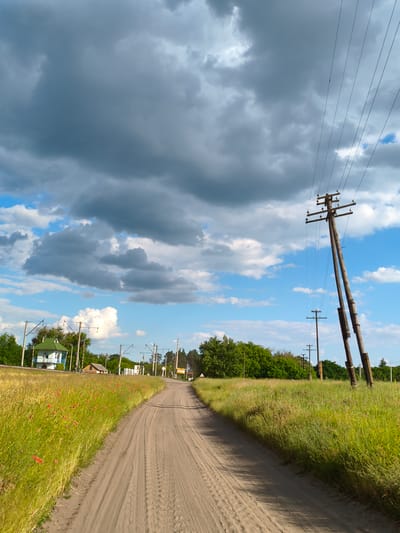 The image depicts a rural scene in Matiashivka, Ukraine, on a cloudy day. A dirt road winds through a grassy field dotted with red poppies and other wildflowers. In the distance, a small white building with a green roof stands beside a railway track. Telephone poles with numerous wires line the road and extend into the background, contributing to the sense of a remote, possibly agricultural, area. The sky is a mix of dark, heavy clouds and patches of bright blue, suggesting an approaching storm or a recent downpour. The overall mood is serene and somewhat dramatic, with the natural elements of the landscape taking center stage. No people are visible, but the tire tracks on the dirt road indicate recent passage. The time of day appears to be late afternoon, given the quality of the light.