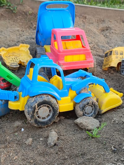 The image shows a collection of colorful plastic toy trucks and other sandbox toys scattered on a sand surface. The primary focus is a blue and yellow front-end loader style toy, covered with sand. Behind it are a pink and yellow truck, and a blue bucket. Other yellow toys are partially visible to the left and right. The toys are set on sand. A few small rocks and some vegetation are present near the base of the front-end loader. There are no people visible in the image. The scene appears to be a children's sandbox or play area. There is no evidence to determine the location or city. The claim that it is in Gabdyukovo, Russia, cannot be verified from the image.