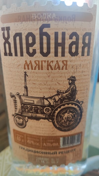 This is a close-up, vertical shot of a bottle of vodka. The label features a vintage-style illustration of a man driving a tractor. Text on the label includes "ВОДКА", "хлебная", "МЯГКАЯ", and "ТРАДИЦИОННЫЙ РЕЦЕПТ". Below the illustration, there are indications of volume (0.5 л), alcohol content (40% об.), and the designation "АЛЬФА". A barcode is also visible. The background is a light, textured paper with faint, dark, thin lines crisscrossing it. The bottle is clear glass, and the overall impression is of a rustic, traditional product. The photograph does not contain any information that places it in the city of Dagomys, Russia.
