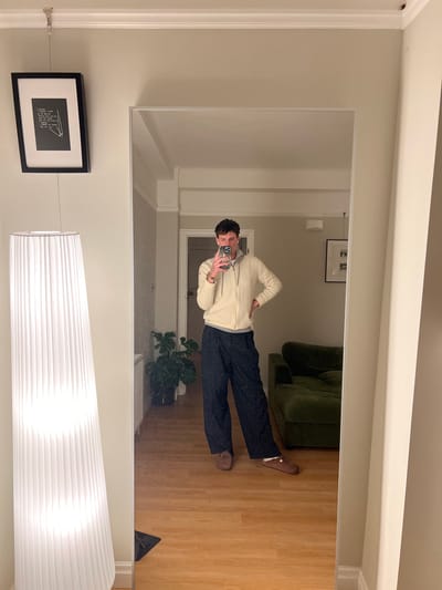 A person stands in front of a mirror, taking a selfie. They wear a cream-colored zip-up sweater, dark blue pants, and brown shoes. The reflection shows a living room setting with a green couch, wooden floor, and a potted plant. Below the mirror, a framed artwork hangs on the wall, and a white pendant lamp is visible.
