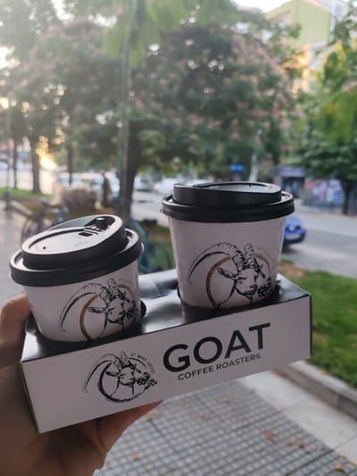 An outdoor, close-up, slightly angled medium shot shows a hand holding a white cardboard coffee cup holder with two white paper coffee cups with black lids, each decorated with a black line drawing of a goat and the text "5th WAVE COFFEE". The holder also features the text "GOAT COFFEE ROASTERS" in large black sans-serif font, with "5th WAVE COFFEE" in a smaller, stylized font. The background is blurred, suggesting an urban environment with trees, buildings, and a street with some vehicles. The lighting indicates it is daytime, possibly late afternoon with sunlight filtering through the trees. The scene is set in Alexandroupolis, Greece.