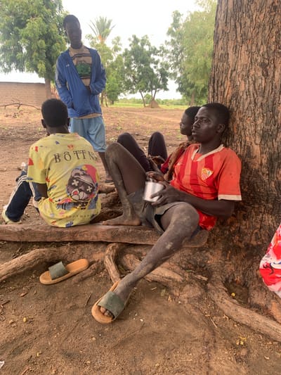 Four young males are seated on the ground under the shade of a large tree, their bodies angled slightly towards the viewer. The scene appears to be outdoors, with the ground covered in dry earth and exposed tree roots. In the background, a few sparse trees and vegetation are visible, suggesting a rural or semi-rural environment. The sky is overcast, indicating potentially cloudy weather.

The individuals are dressed casually. One person in the foreground wears a yellow t-shirt with a cartoon graphic and the text "BÔTTN" and "OIPUSHAYU" printed on it. Another person is wearing a red and white striped t-shirt and dark shorts, holding a metal cup. A third person is in a blue jacket and light-colored pants, and the fourth is mostly obscured but appears to be wearing dark pants and a t-shirt. Their postures suggest they are resting or seeking shelter from the sun.

The time of day appears to be daytime, given the light conditions and the presence of shade. The ground is dry and dusty, with some small pebbles and leaves scattered around. The roots of the large tree are prominently featured, intertwining with the ground. Two pairs of sandals are visible on the ground near the seated individuals. The overall impression is of a simple, informal gathering in a natural setting.