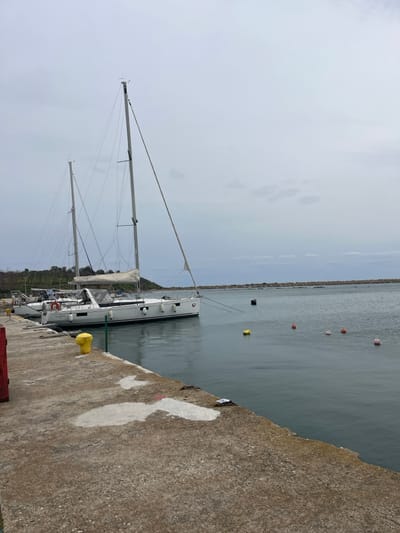 This image depicts a marina in Limenas Avdiron, Greece, under an overcast sky.
In the foreground, a weathered concrete pier extends from the lower left to the lower right, featuring irregular white patches and small scattered debris on its surface. A yellow cylindrical bollard is prominently located on the pier, slightly to the left of the center, with a red vertical structure partially visible on the far left edge.
The midground showcases calm, dark grey-green water where several sailboats are moored. The primary subject is a large, white sailing yacht with a grey stripe along its hull, equipped with two masts and multiple black cylindrical fenders attached to its starboard side. Mooring lines extend from the yacht. Floating in the water are one yellow cylindrical buoy, three red spherical buoys, and one black cylindrical buoy, spaced across the visible water surface.
The background consists of a low, green-vegetated landmass or hill on the left. The horizon is marked by a long, low-lying grey stone breakwater extending horizontally across the mid-distance. The sky above is uniformly overcast, presenting a light grey color with no distinct cloud formations. No people are present in the scene.