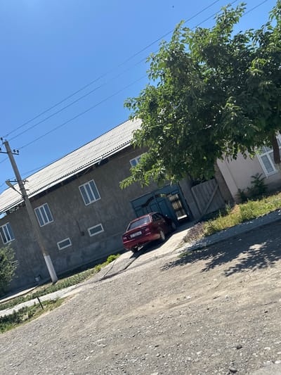 In the city of Тынчтык, Kyrgyzstan, a tilted, low-angle outdoor shot captures a residential scene under a clear blue sky. The foreground is a dirt and gravel road, with patches of green grass and shadows from trees suggesting a sunny day. To the left, a gray, two-story building with a corrugated metal roof dominates the frame. The building has several rectangular windows with white frames. A utility pole with wires is visible near the left edge of the building. In front of the building, a red sedan with a license plate is parked near a teal-colored metal gate. The car is facing away from the viewer and towards the gate. To the right of the building, part of another house is visible, also with white-framed windows, and a large tree with dense green foliage stands between the two structures, casting shadows on the ground. The overall impression is of a quiet, everyday street in a residential area.