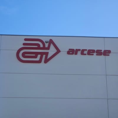 The photograph shows the "arcese" logo on the side of a building. The logo is red and consists of a stylized letter "G" merging into an arrow pointing to the right, followed by the word "arcese" in red block letters. The building's exterior is white with horizontal panel lines. Above the logo is a security light. The background is a clear blue sky. The location is Alcalá de Henares, Spain.