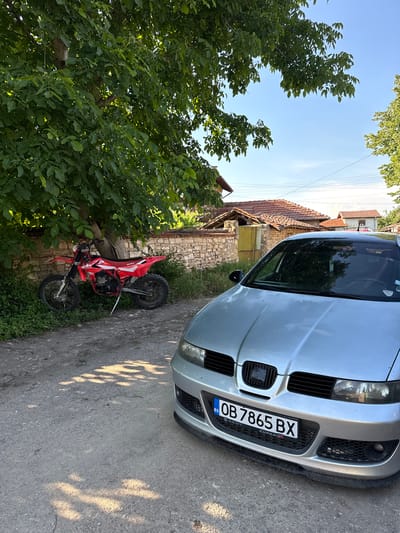 In Bratya Daskalovi, Bulgaria, a silver car is parked on a dirt road in front of a stone wall. A red dirt bike is parked to the left of the car, under a tree with green leaves. The car has a license plate that reads "OB 7865 BX". The background shows several houses with red tile roofs and a clear blue sky. The sun is shining, casting shadows on the road.