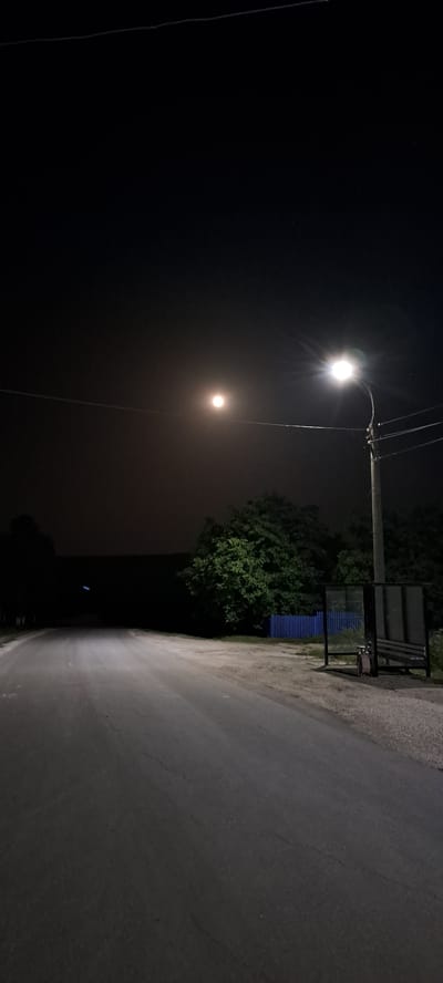 The image depicts a nighttime scene on a rural road, likely in Rădoaia, Moldova. The road is paved and has visible cracks. To the right, there is a bus stop with a bench and a shelter. A streetlamp illuminates the scene, casting a bright glow and creating a starburst effect. In the background, there are trees and a blue fence. The sky is dark with a hazy moon visible. There are no people or animals present in the image. The overall impression is one of a quiet, dimly lit rural setting at night.