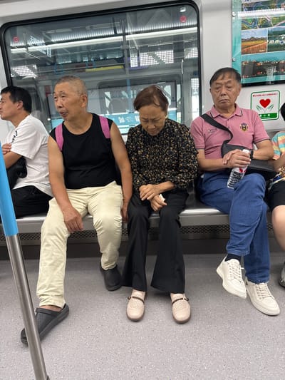 The image shows a group of people seated on a train, likely a subway, in Shuangfu, China. Four people are visible: an elderly man in a pink polo shirt holding a water bottle, a woman in a floral print blouse, a man in a black tank top with pink trim, and another man in a white t-shirt. The scene suggests a casual and possibly mundane moment in urban public transit. The presence of a "courtesy seat" sign indicates consideration for those in need of seating.