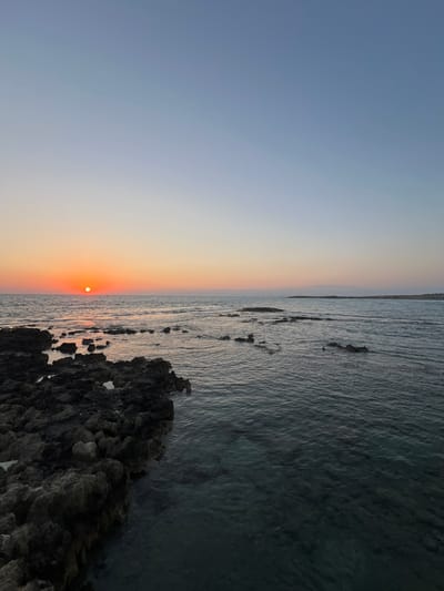 This media file captures a serene sunset over the sea. The scene is divided horizontally, with the left side dominated by a gradient of the sky transitioning from a soft blue to a warm orange hue, indicating the setting sun. The sun itself is a small, bright orange disc positioned just above the horizon line, casting a subtle glow.
The right side of the image depicts the tranquil sea. The water is a deep, cool tone, with gentle ripples suggesting a calm environment. Scattered rocks and possibly shallow coral formations are visible beneath the water's surface, breaking the expanse of the sea. In the foreground, a cluster of dark, textured rocks juts out into the water, adding a sense of grounding to the scene.
The overall mood conveyed is peaceful and contemplative, characteristic of a sunset by the sea. The time of day is clearly evening, as the sun is low on the horizon. The weather appears clear, with no visible clouds. The location is identified as Akrotiri, Sovereign Base Areas of Akrotiri and Dhekelia, suggesting a coastal setting in Cyprus. There is no visible text in the image. The composition, with its stark horizontal division between sky and sea, emphasizes the vastness and beauty of nature.