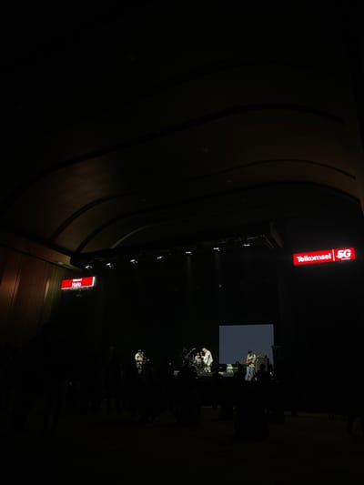 The image shows a dimly lit indoor space in Salembaran, Indonesia. A stage with musicians performing is visible under spotlights, with a screen behind them. Two large, illuminated signs for "Telkomsel 5G" and "Telkomsel Halo" are positioned above the stage. The walls have a curved, textured design. The overall environment is dark, with only the stage and signs brightly illuminated.