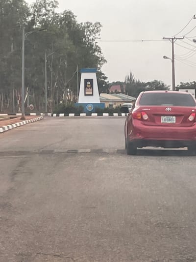 The image shows a red Toyota Corolla parked on the side of a road in Afaka, Nigeria. The vehicle's license plate reads "YAB-844GE." A building with a white and blue sign is visible in the background. Trees line the road, and overhead utility lines are present. The sky is overcast.