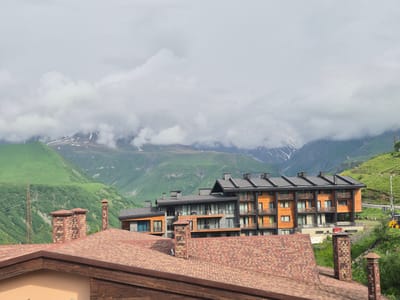 A wide shot shows a modern, multi-story hotel building with orange and gray facades, situated in a mountainous region with green slopes and snow-capped peaks under a cloudy sky. In the foreground, the reddish-brown tiled roofs of two buildings are visible, with decorative brick chimneys. A power line tower stands to the left, and a yellow construction vehicle is parked on the right. The scene is likely Gudauri, Georgia, a popular ski resort town known for its mountainous terrain.